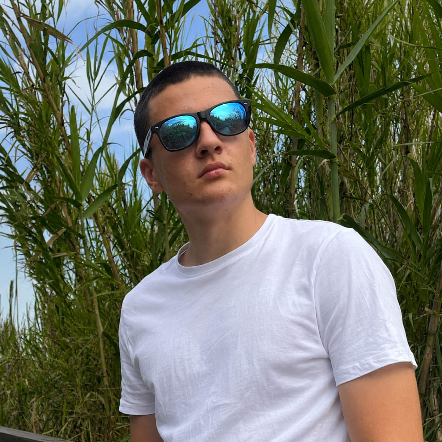 Man wearing wooden sunglasses and a white t-shirt standing in front of tall green plants.